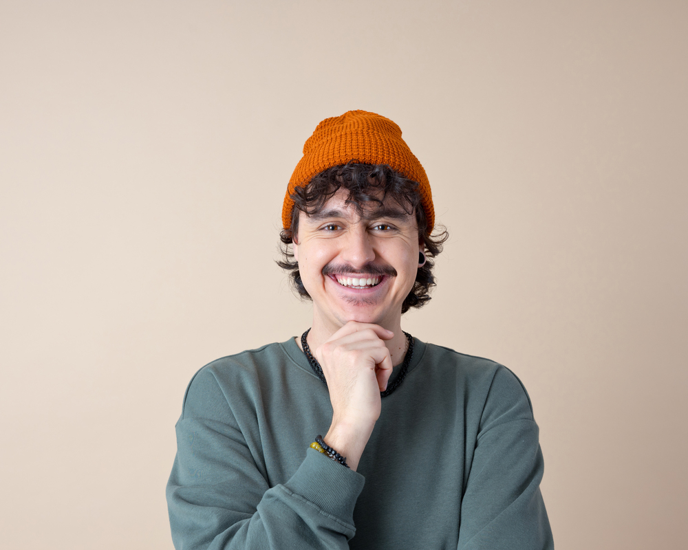 Ein lachender Mann mut Schnurbart wirbt für Männergesundheitustache wearing khaki blouse and orange beanie smiling at camera. Studio shot, beige background.